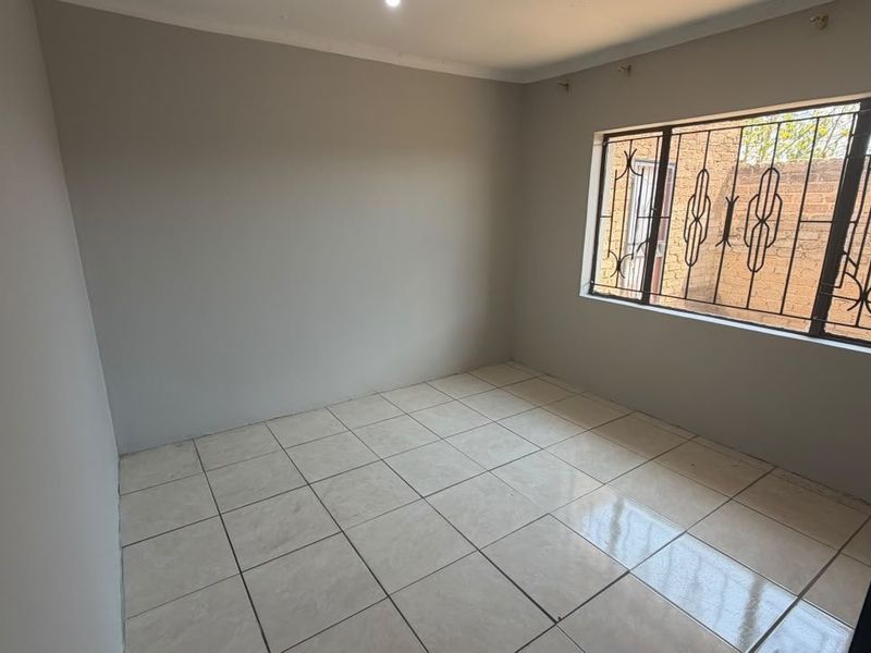 A clean, empty bedroom featuring neutral wall paint, tiled flooring, and a window fitted with security bars.