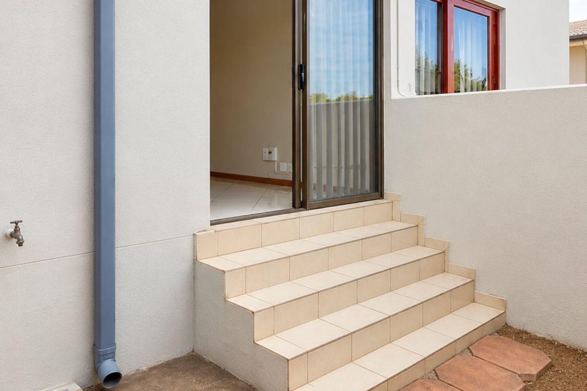 Rear exterior view of the townhouse featuring tiled steps leading to a sliding glass door, paved walkway, and functional drainage infrastructure.