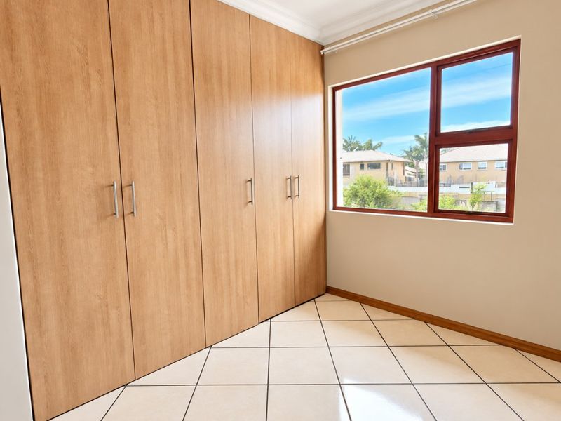 A bright, empty bedroom featuring light wood-finish built-in cupboards, neutral tiled flooring, and a window with a view of the surrounding neighborhood.