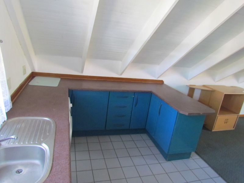 A functional kitchen area featuring blue cabinetry, laminate countertops, a stainless steel sink, and exposed ceiling rafters in an attic-style layout.