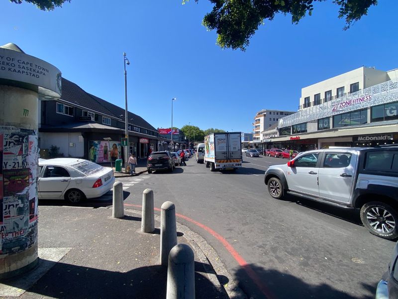 A street-level view of a busy commercial area featuring storefronts, road infrastructure, and concrete bollards along the sidewalk.