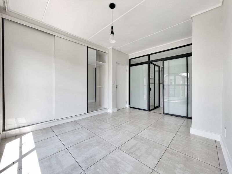 A modern, bright bedroom featuring large-format floor tiles, a sleek built-in sliding wardrobe, and a contemporary black-framed glass partition.