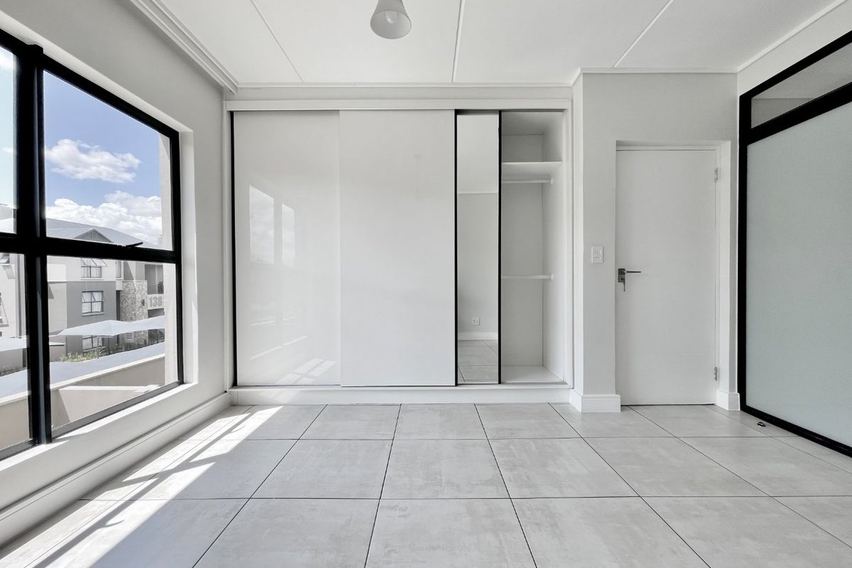 A bright, modern bedroom featuring large windows, neutral tiled flooring, and a sleek built-in sliding wardrobe with integrated shelving.