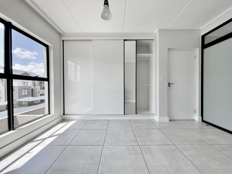 A bright, modern bedroom featuring large windows, neutral tiled flooring, and a sleek built-in sliding wardrobe with integrated shelving.