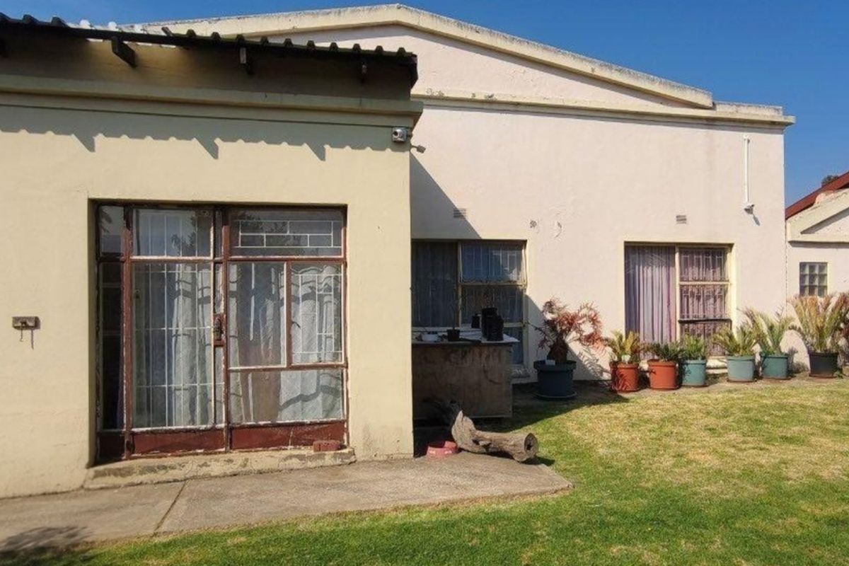 Rear view of the property featuring a concrete patio, steel-framed windows, and security gates, set against a garden area with visible exterior maintenance required.