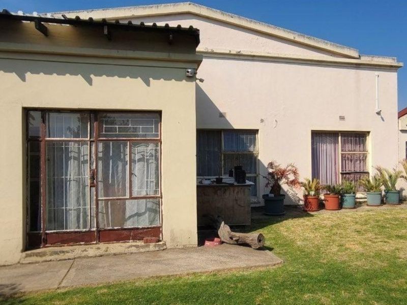 Rear view of the property featuring a concrete patio, steel-framed windows, and security gates, set against a garden area with visible exterior maintenance required.