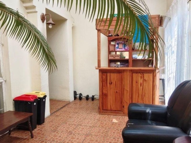 A casual indoor bar area featuring a built-in wooden counter, tiled flooring, and natural light from large windows with security bars.