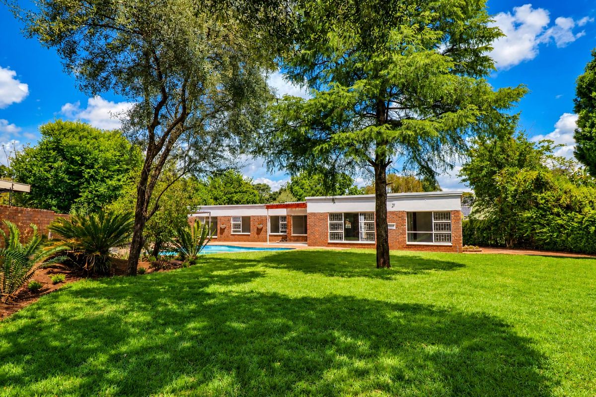 Rear view of the property featuring a spacious lawn, mature trees, a swimming pool, and a brick facade with large sliding glass doors.