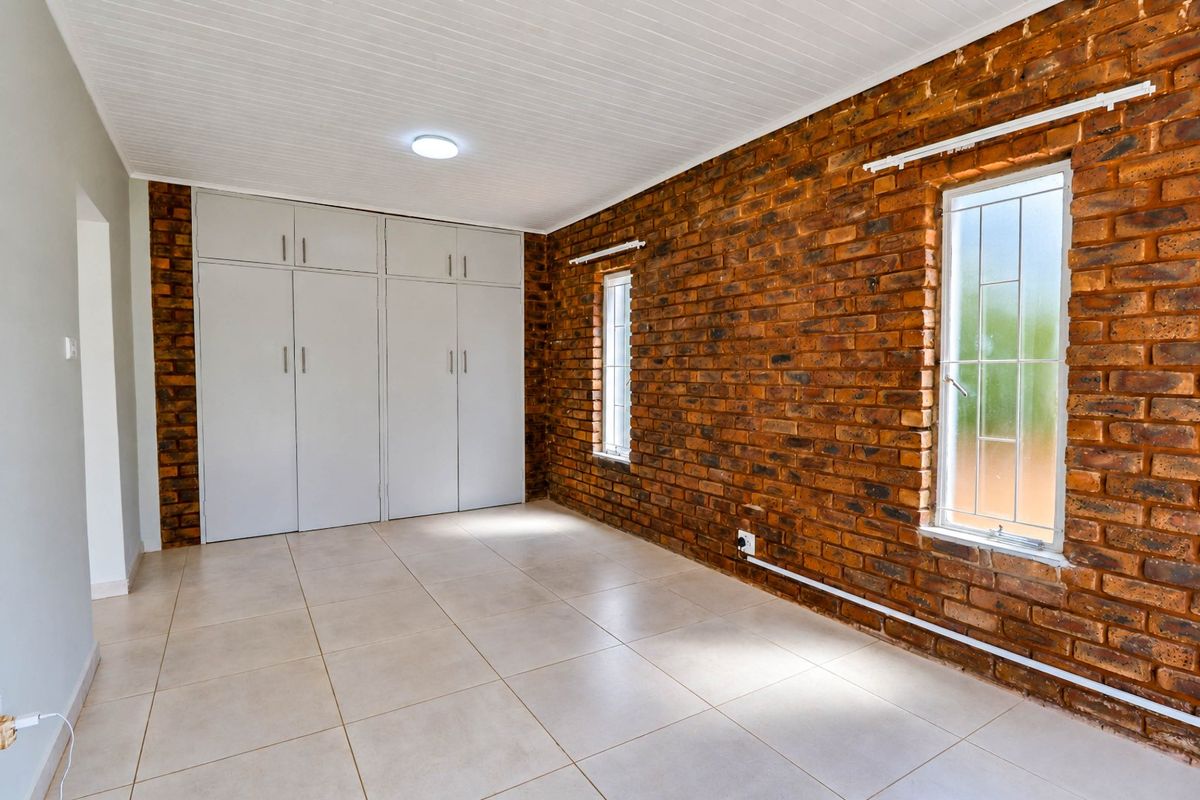 A bright, empty bedroom featuring a distinctive exposed brick wall, tiled flooring, and ample built-in cupboard storage space.