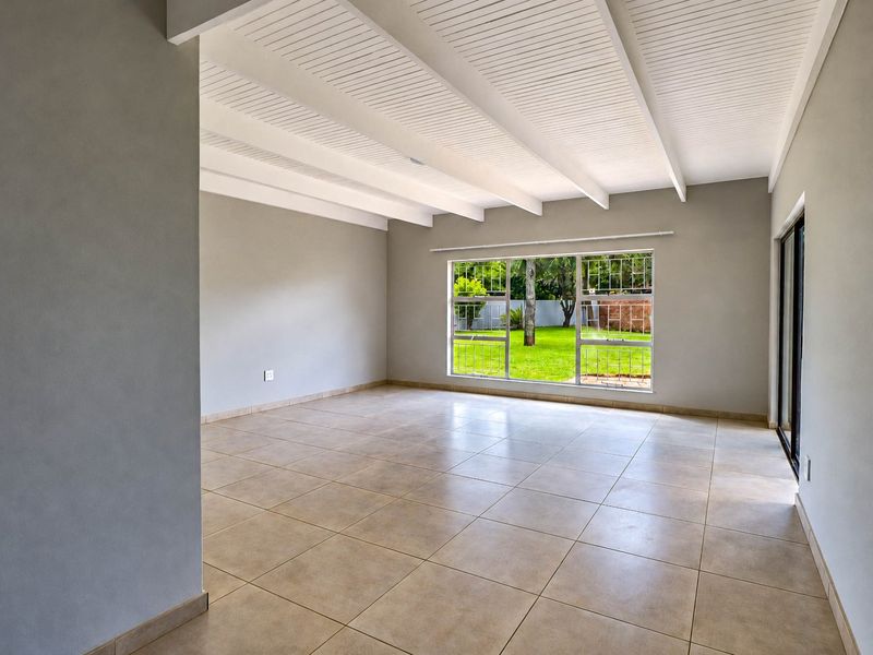 A bright, empty living room featuring tiled floors, an exposed beam ceiling, and large windows overlooking the garden.