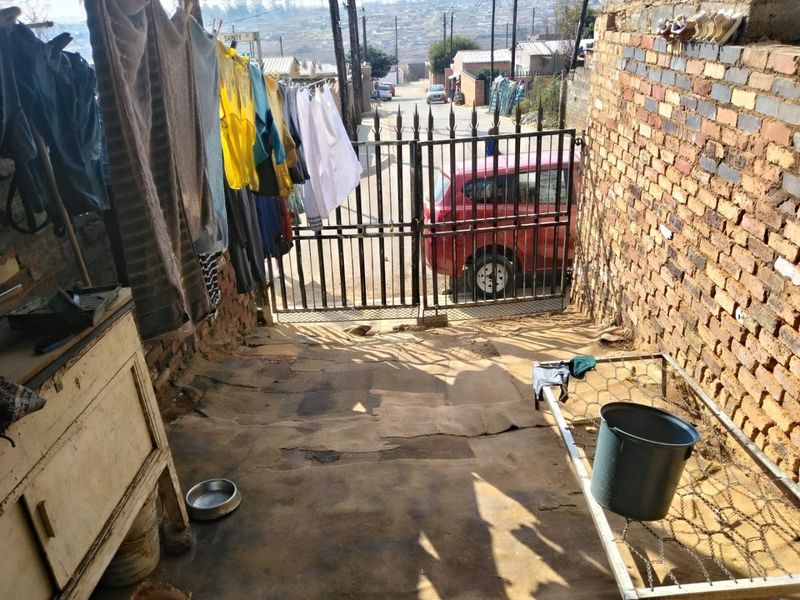 A narrow exterior entrance area featuring a metal security gate, exposed brick boundary walls, and a concrete driveway surface.
