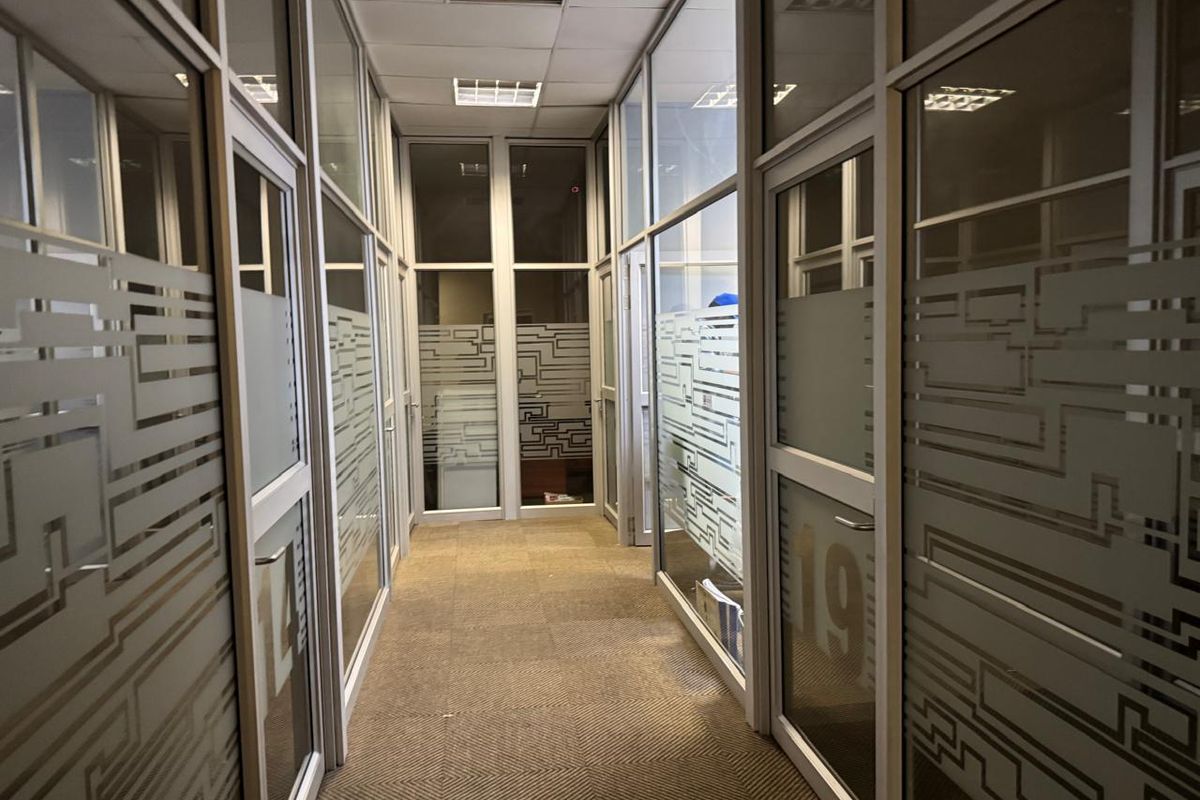 A commercial office hallway featuring glass partition walls with frosted privacy film, carpeted flooring, and a suspended ceiling with recessed lighting.