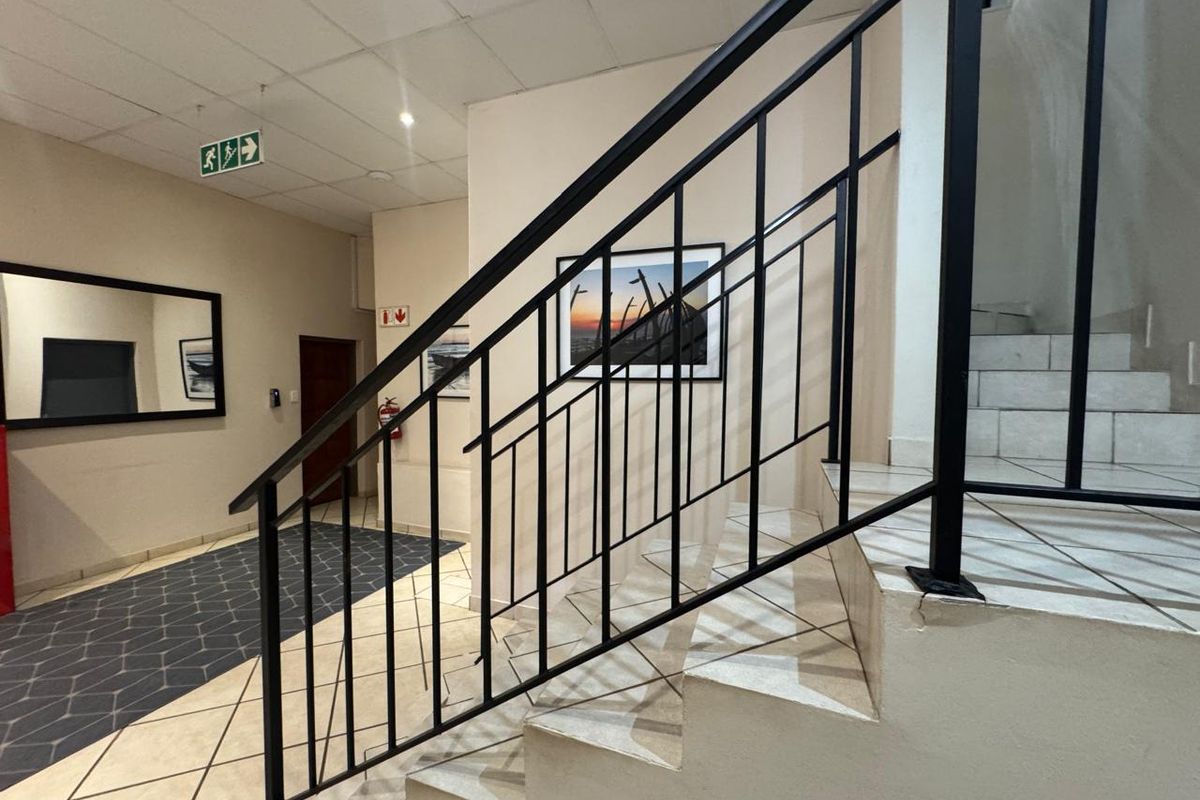 A commercial hallway featuring a staircase with a black metal balustrade, tiled flooring, and a suspended ceiling with emergency signage.