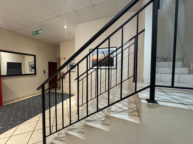 A commercial hallway featuring a staircase with a black metal balustrade, tiled flooring, and a suspended ceiling with emergency signage.