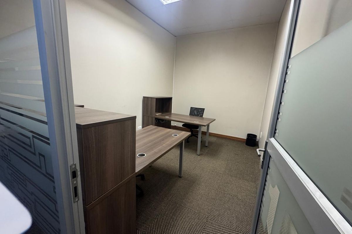 A functional office space featuring carpeted flooring, a suspended ceiling with recessed lighting, and a modern frosted glass entry door.