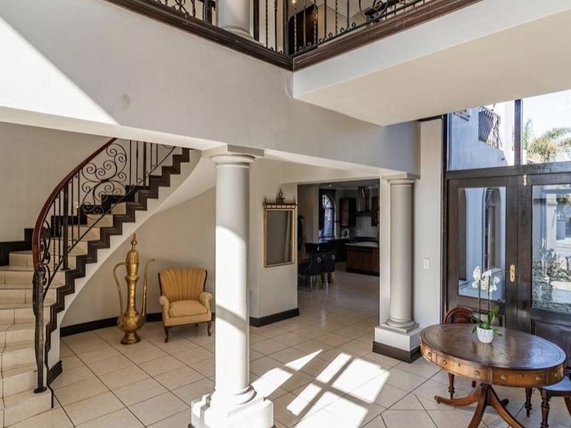 Grand double-volume entrance hall featuring a sweeping curved staircase with wrought iron balustrade, tiled flooring, and classical columns leading to an open-plan living area.