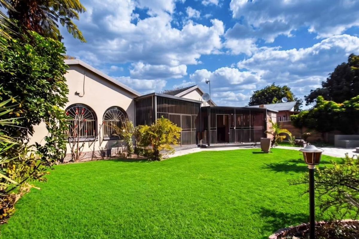 A well-maintained rear garden featuring a lush lawn, paved patio area, and an enclosed patio structure with arched windows and security features.