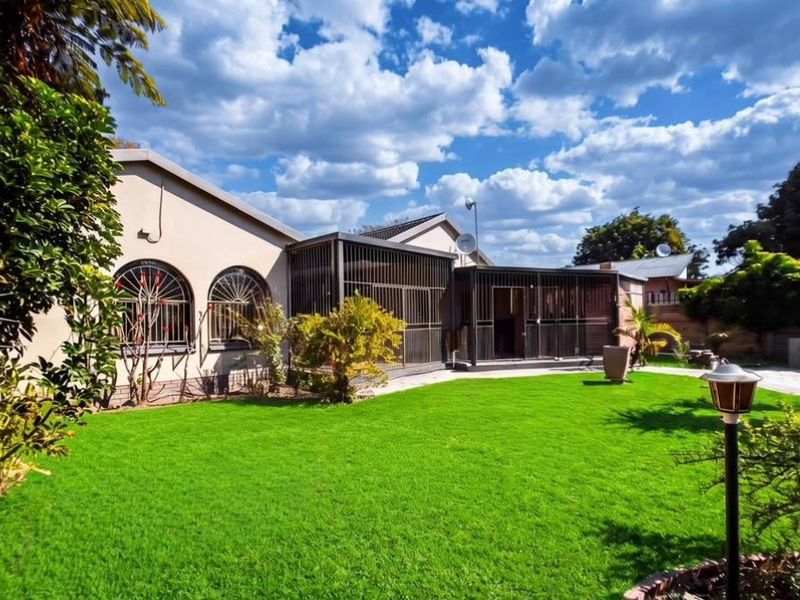 A well-maintained rear garden featuring a lush lawn, paved patio area, and an enclosed patio structure with arched windows and security features.