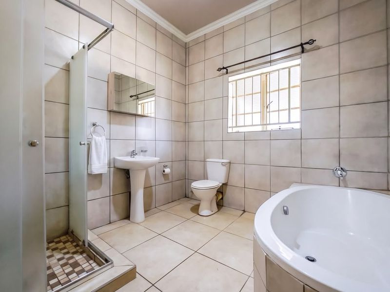 A spacious, tiled bathroom featuring a corner bathtub, separate shower enclosure, pedestal basin, and toilet, with natural light from a window.