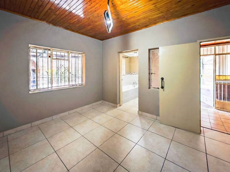 An empty bedroom featuring tiled flooring and a timber tongue and groove ceiling, with direct access to an adjoining bathroom and hallway.