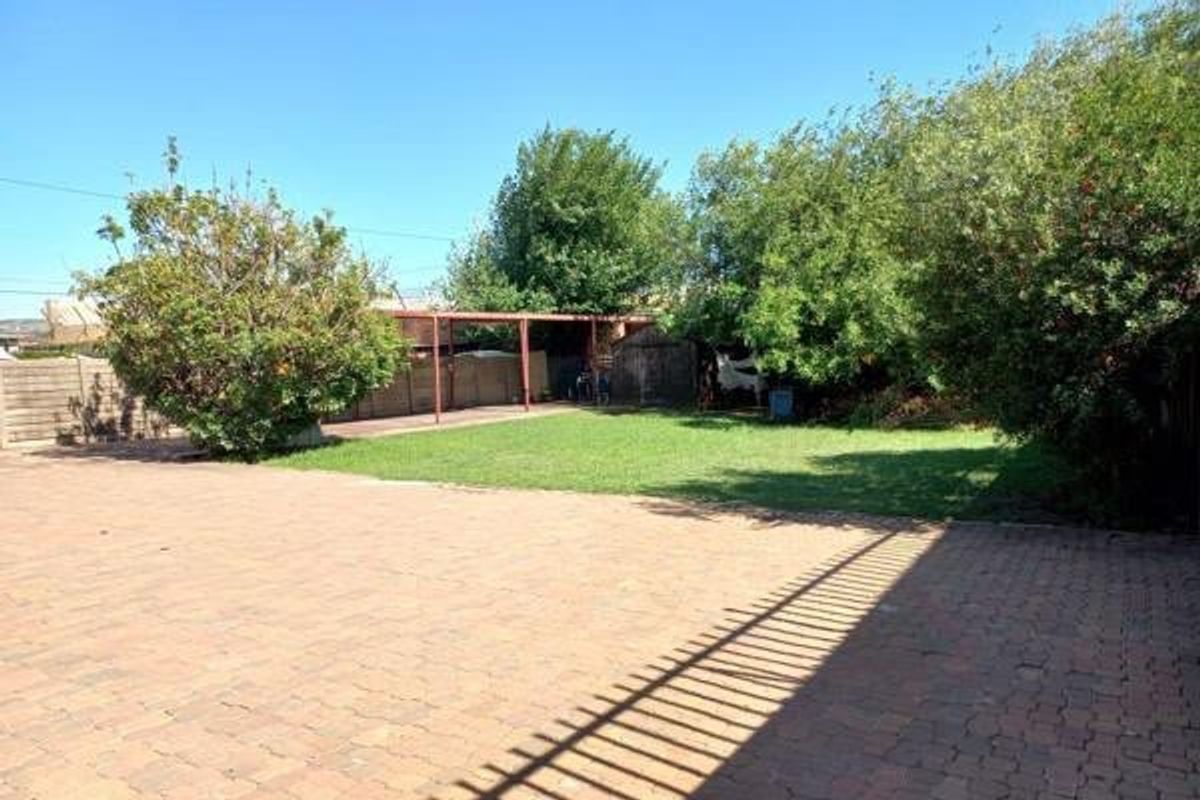 Spacious rear garden featuring a large paved driveway, a well-maintained lawn, and a covered patio area surrounded by mature trees and perimeter fencing.