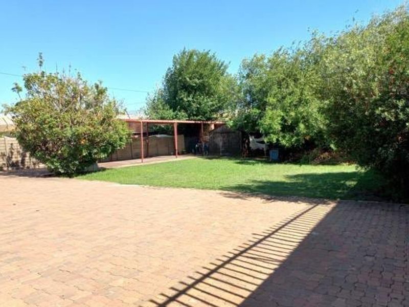 Spacious rear garden featuring a large paved driveway, a well-maintained lawn, and a covered patio area surrounded by mature trees and perimeter fencing.