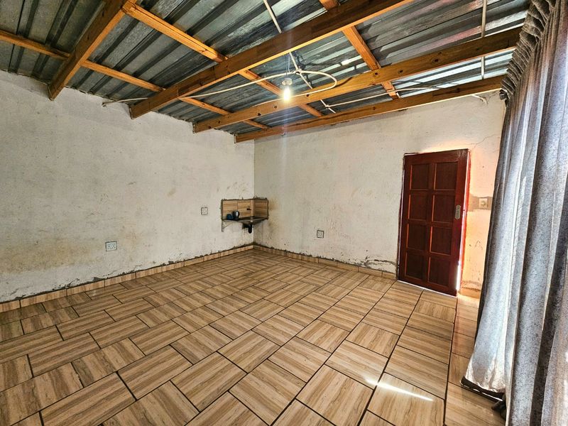 An empty living area featuring exposed timber roof trusses, corrugated metal ceiling, and ceramic tiled flooring, requiring cosmetic attention to wall finishes.