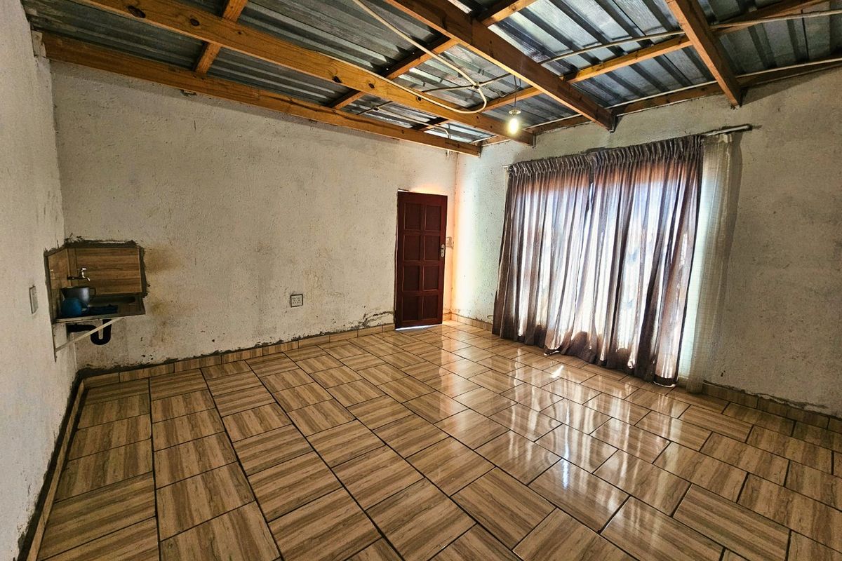 An empty living area featuring tiled flooring, exposed timber roof trusses, and a wall-mounted basin, requiring cosmetic renovation and wall finishing.