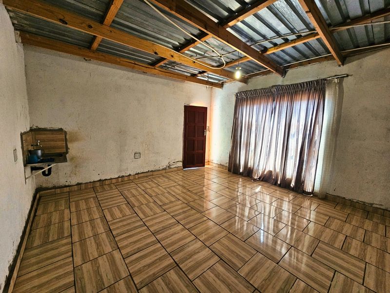 An empty living area featuring tiled flooring, exposed timber roof trusses, and a wall-mounted basin, requiring cosmetic renovation and wall finishing.