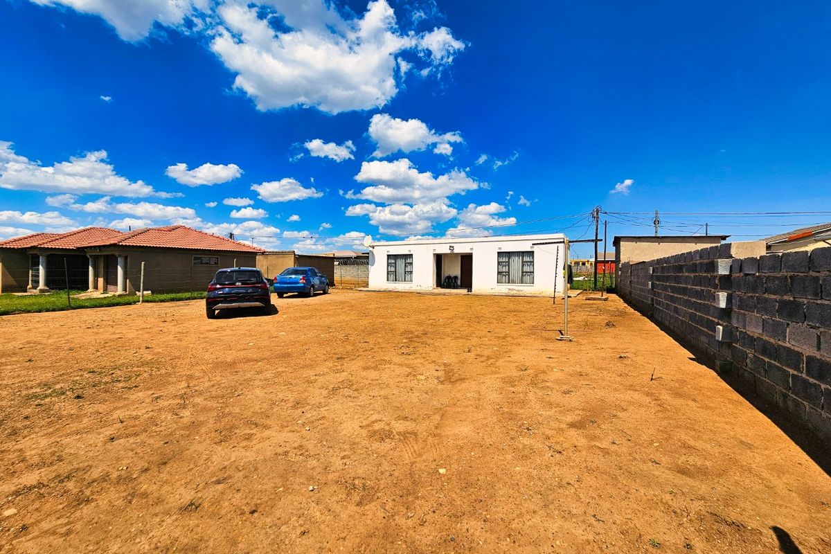 Front view of a single-story residential house featuring a large unpaved yard and a concrete block boundary wall.