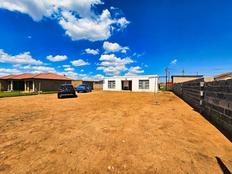 Front view of a single-story residential house featuring a large unpaved yard and a concrete block boundary wall.