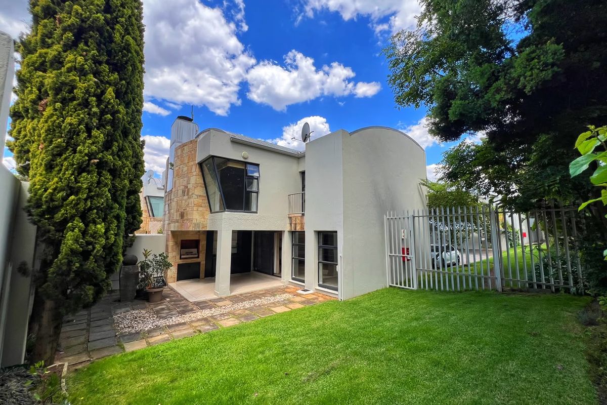 Rear view of the property featuring a paved patio area, built-in braai, stone facade accents, and a well-maintained garden with security fencing.