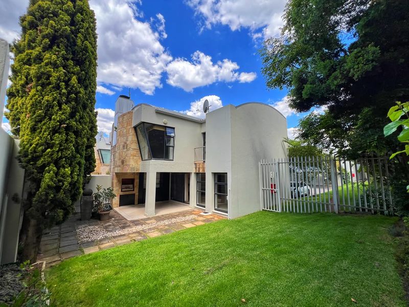 Rear view of the property featuring a paved patio area, built-in braai, stone facade accents, and a well-maintained garden with security fencing.