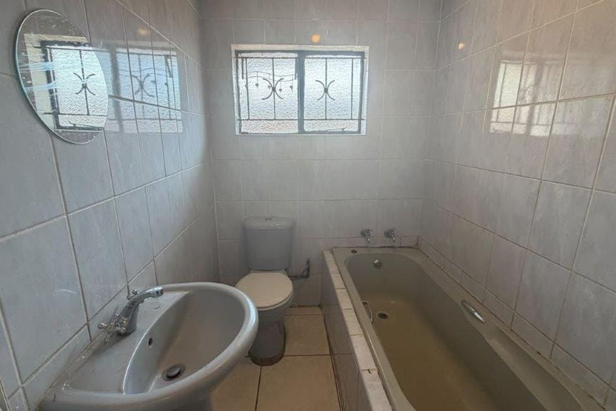 A functional bathroom featuring a pedestal basin, toilet, and built-in bathtub with full-height wall tiling and a window with security bars.
