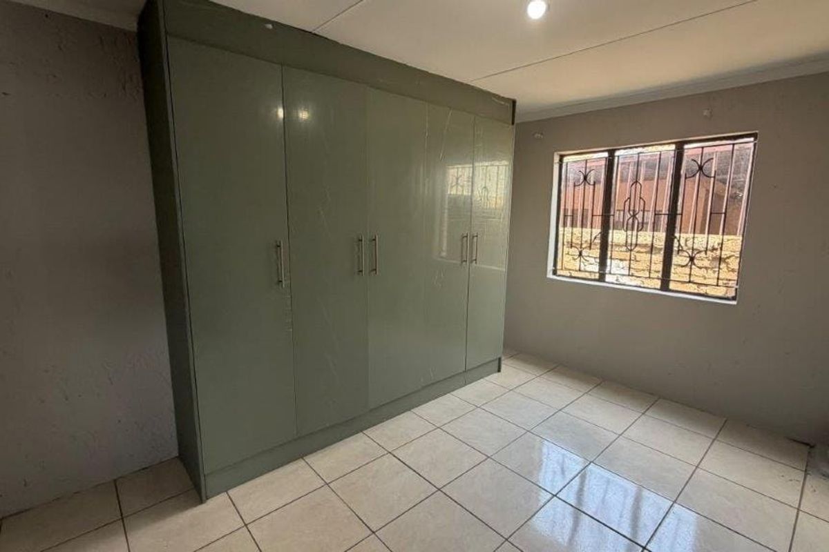 A clean, empty bedroom featuring neutral tiled flooring and large built-in cupboards with modern grey doors and security bars on the window.