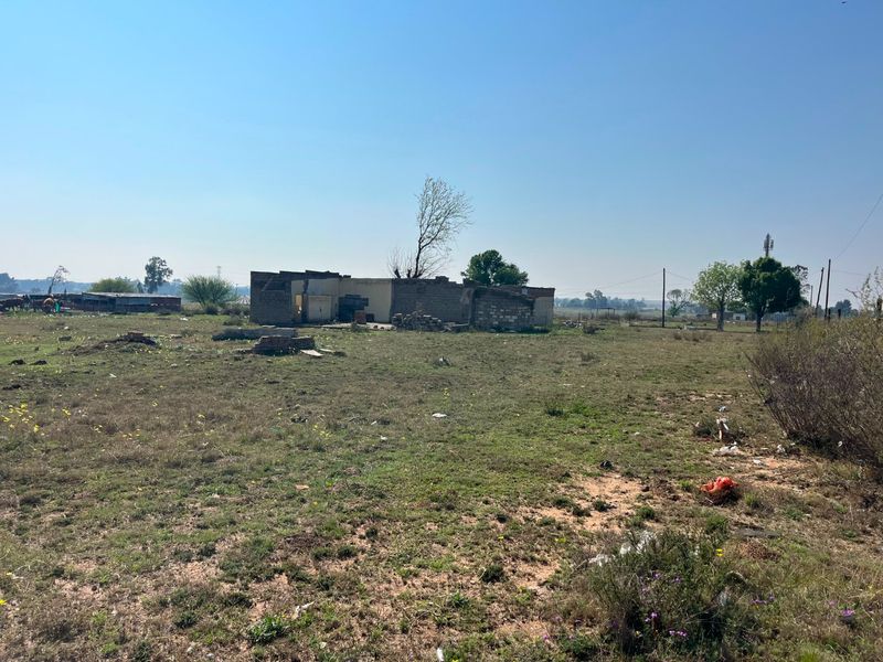 A view of a dilapidated, partially demolished brick structure situated on a large, undeveloped rural farm plot.
