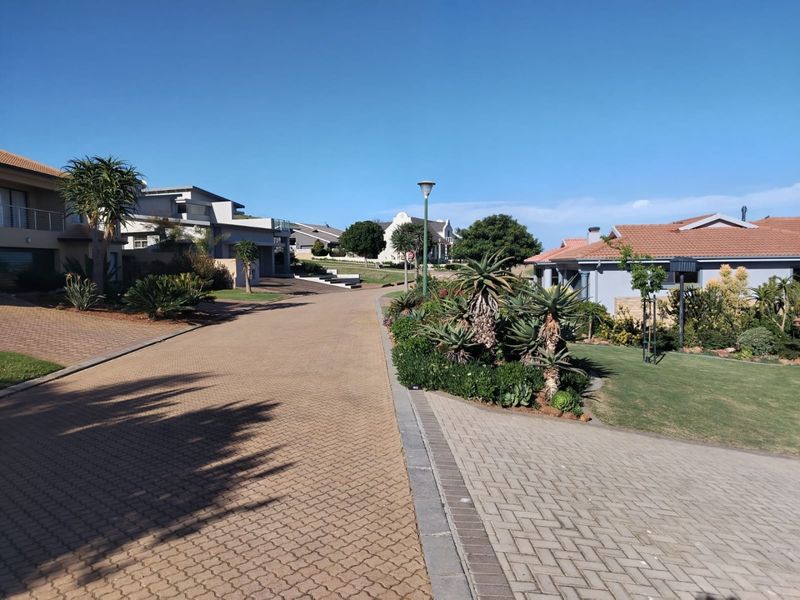 A well-maintained residential street scene featuring paved driveways, established garden landscaping, and modern housing architecture under a clear blue sky.