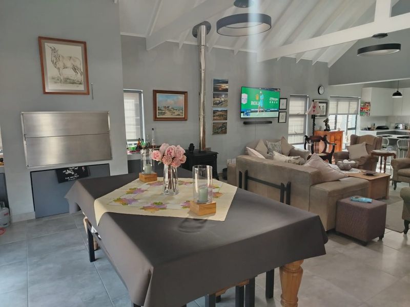 Spacious open-plan living area featuring a vaulted ceiling with exposed beams, a built-in indoor braai, and a cozy fireplace, seamlessly connecting to the kitchen.