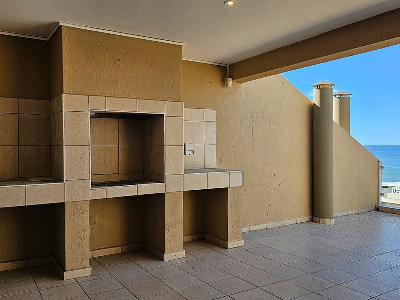 Spacious covered balcony featuring a built-in braai, tiled flooring, and a glass balustrade with panoramic ocean views.
