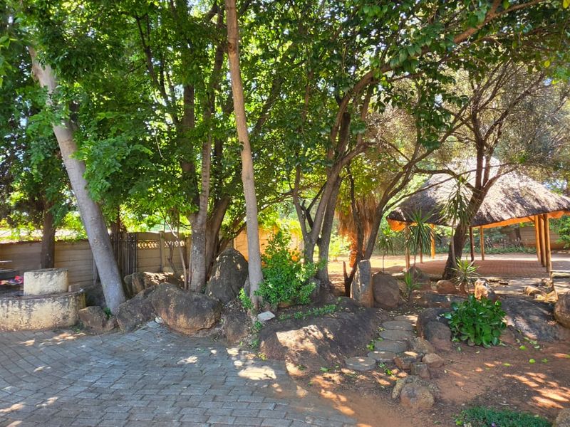 A spacious rear garden featuring mature trees, a brick-paved patio area, a built-in braai, and a traditional thatched lapa for outdoor entertainment.