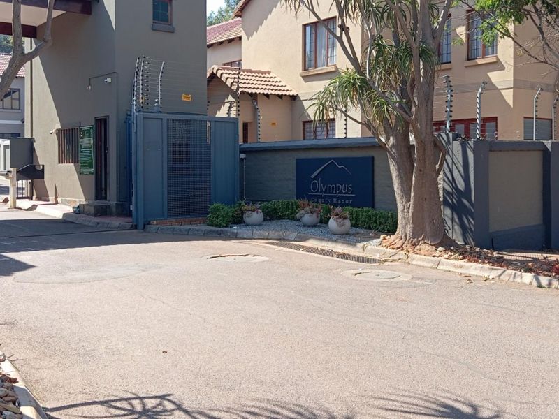 Entrance view of Olympus Country Manor featuring a secure gated access point, boundary walls with electric fencing, and a paved driveway.