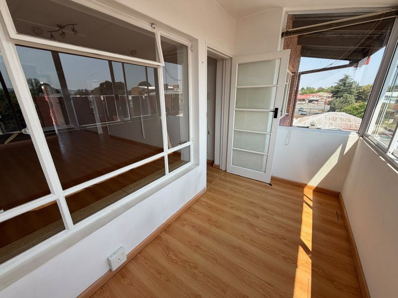 An enclosed balcony or sunroom featuring laminate flooring, large windows providing natural light, and a glass panel door leading to the interior.