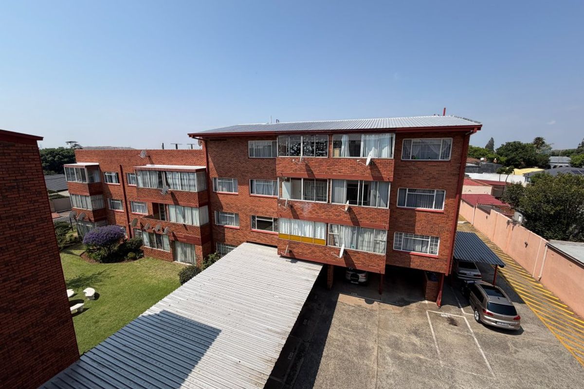 Exterior view of a multi-story brick apartment complex featuring a communal garden, paved parking area, and covered carports.
