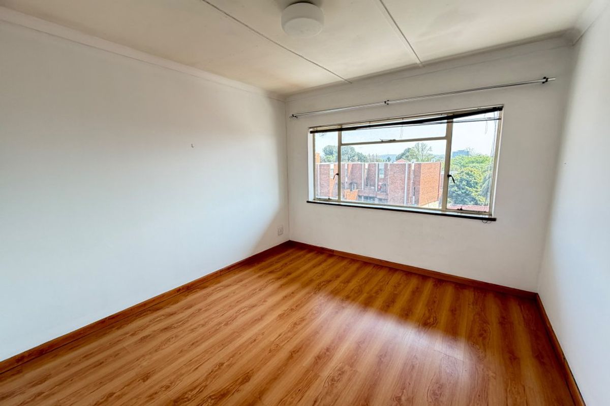 A bright, empty bedroom featuring laminate flooring, a large window with a curtain rod, and a ceiling-mounted light fixture.