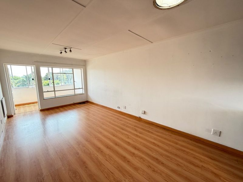 An empty, sunlit living room featuring laminate flooring, neutral walls, and large aluminium-framed windows with access to an adjoining balcony.