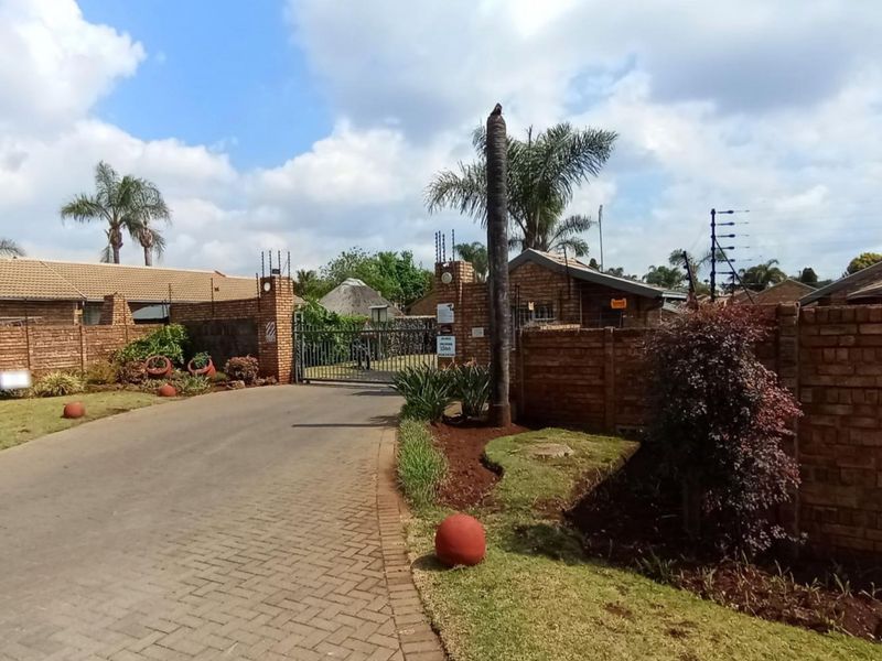 Street view of a townhouse complex entrance featuring brick boundary walls, electric fencing, and a paved driveway leading to a security gate.