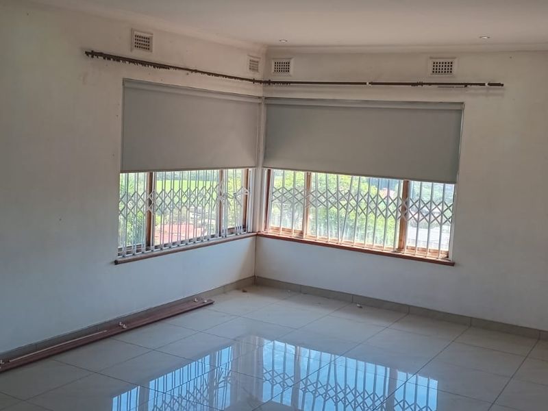 An empty living room featuring large corner windows with security gates, roller blinds, and polished tiled flooring throughout.