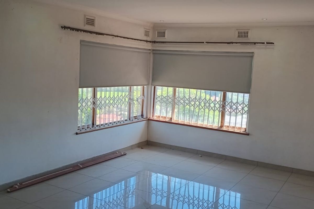 An empty living room featuring large corner windows with security gates, roller blinds, and polished tiled flooring throughout.