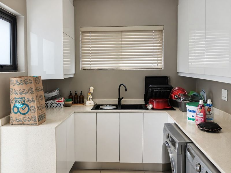 A modern, compact laundry room featuring white high-gloss cabinetry, stone countertops, a double basin sink with a black mixer tap, and tiled flooring.