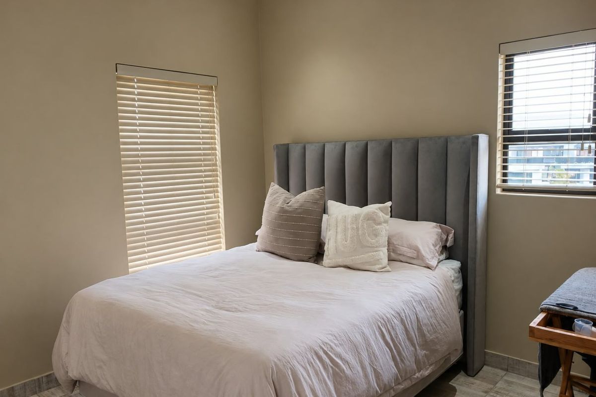 A well-maintained bedroom featuring neutral wall tones, tiled flooring, and dual windows fitted with venetian blinds.